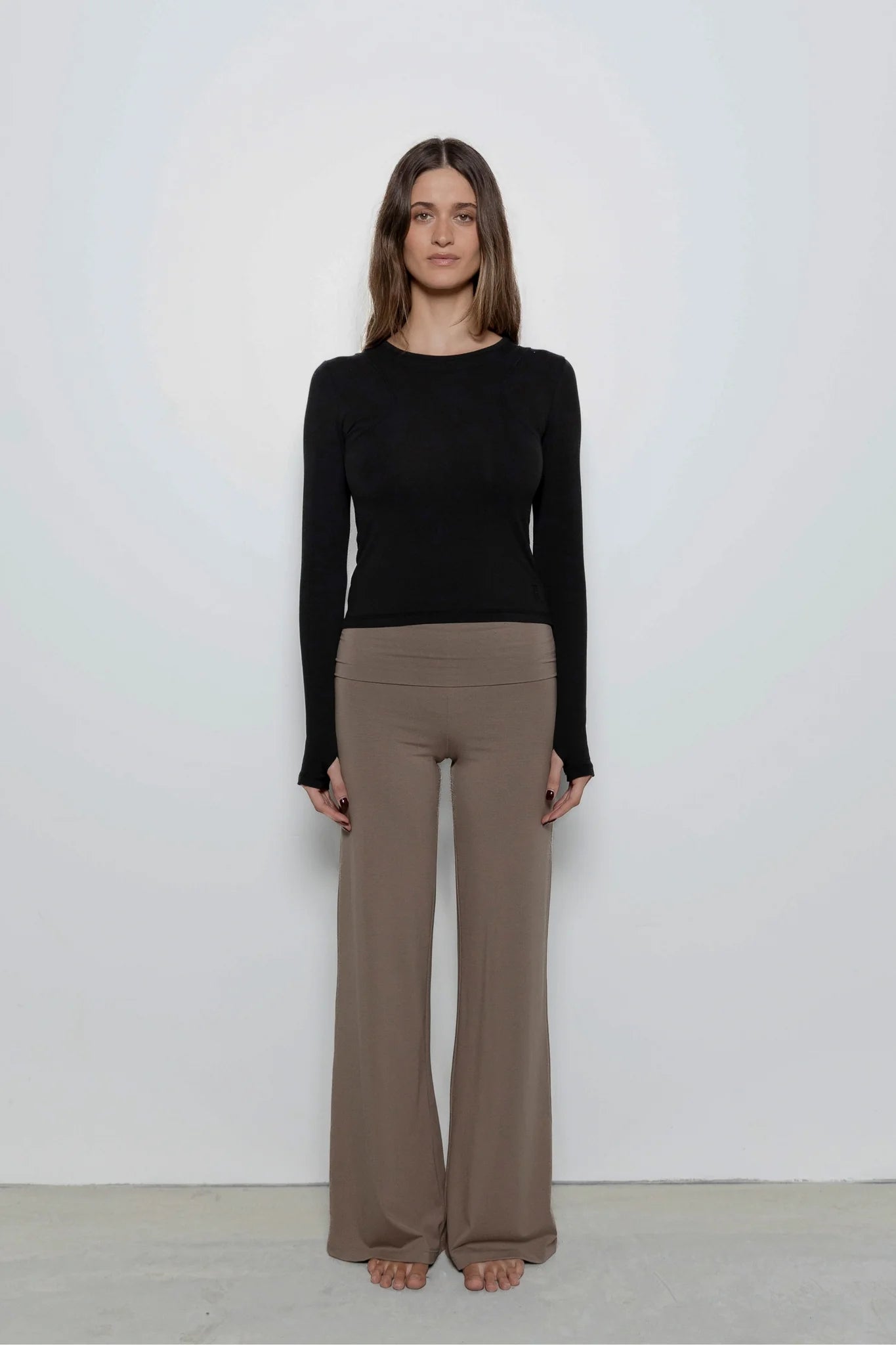 A woman stands facing forward in a studio wearing a fitted black long-sleeve top and loose taupe-brown wide-leg pants against a plain light gray wall.